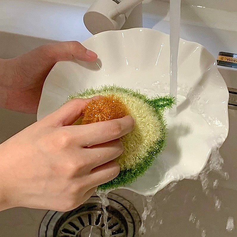 Hand washing a green avocado shape sponge under running water in a sink.
